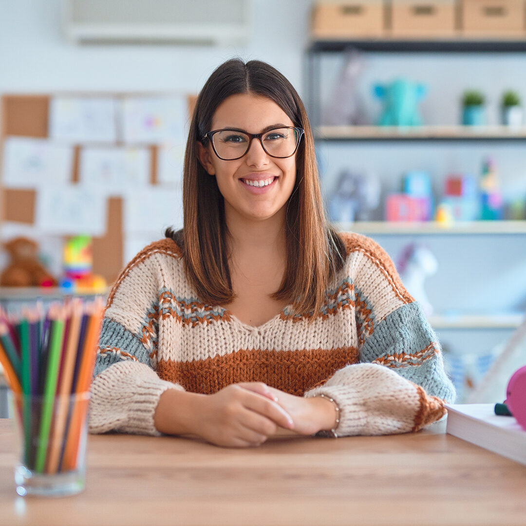 Young beautiful teacher woman wearing sweater and glasses sitting on desk at kindergarten with a happy and cool smile on face. Lucky person.