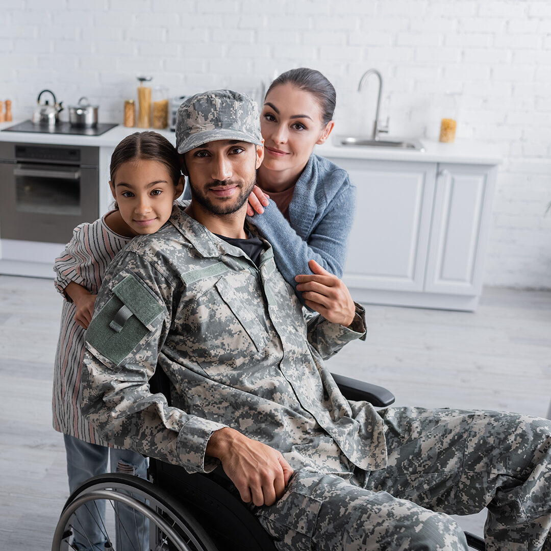 Man in military uniform and wheelchair looking at camera near family at home