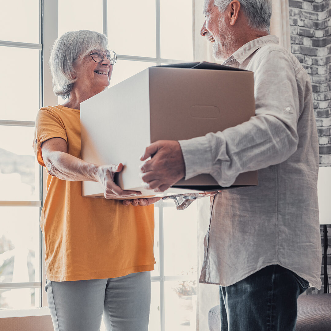 Mature couple moving into new apartment, carrying cardboard boxes into empty room with potted plants. Real estate property buying, relocation, new home concept. Rear view